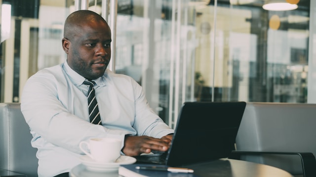 man in white shirt and tie in office working on laptop with coffee cup