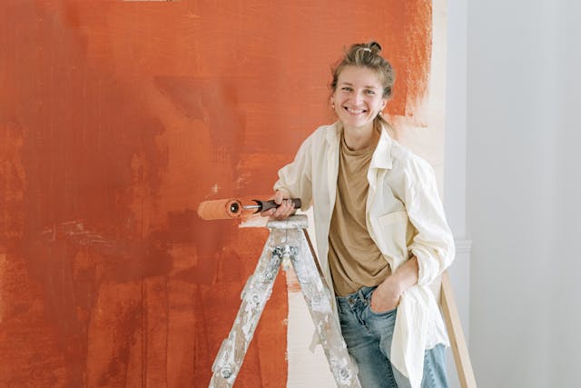pretty blonde woman smiling standing on ladder with roller and wall being painted rust color