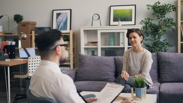 woman sitting on a couch talking to a man holding a clipboard sitting across from her
