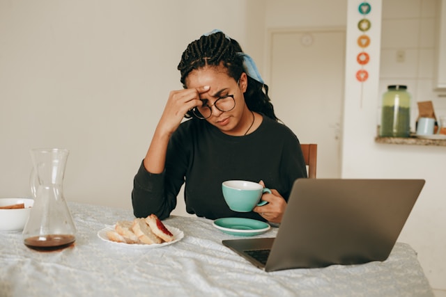 black woman in black shirt holding coffee at table with laptop