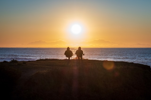 couple sitting on edge of coastline as sun sets in the horizon