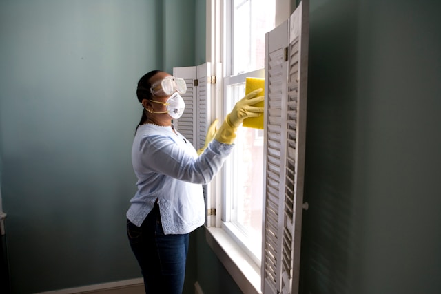 woman in whote short and jeans wearing mask with yellow gloves cleaning window with a yellow sponge