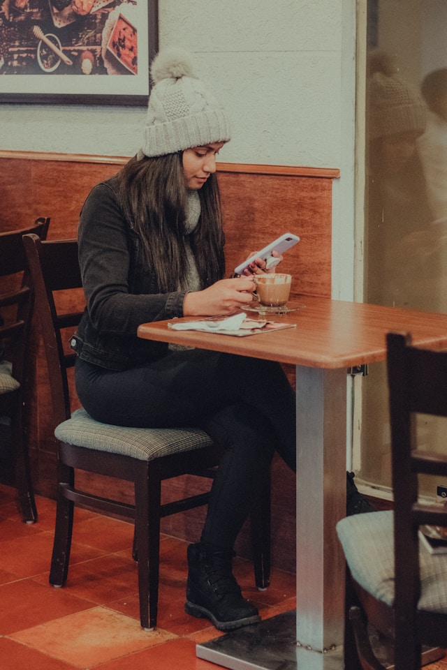 pretty woman with long dark hair wearing knit hat looking at her smartphone while sitting on chair by table