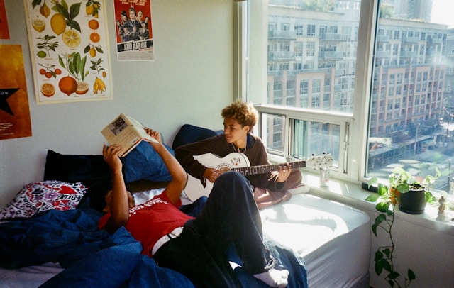 two guys sitting on bed one playing guitar