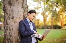 Asian man in blie blazer leaning against a tree and hold an open laptop