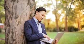 Asian man in blie blazer leaning against a tree and hold an open laptop