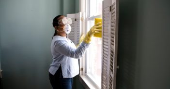 woman in whote short and jeans wearing mask with yellow gloves cleaning window with a yellow sponge