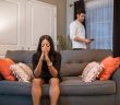 woman in black dress sitting on couch with hands to her face with man in white shirt standing behind couch looking at his phone