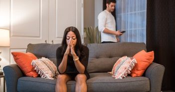woman in black dress sitting on couch with hands to her face with man in white shirt standing behind couch looking at his phone