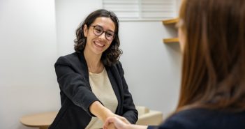 woman with dark hair and glasses in jacket and blouse shaking hands with woman across the table