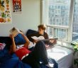 two guys sitting on bed one playing guitar