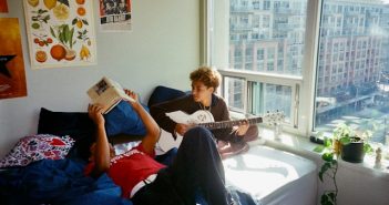 two guys sitting on bed one playing guitar