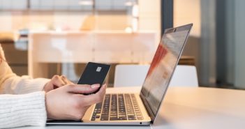 Person seated at a table using a laptop and holding a credit card while shopping online at home